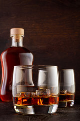 A glass of whiskey and ice on the wooden table. In the background, a glass of whiskey with ice and a full bottle of drink.