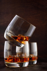 Two glasses of whisky and ice on the wooden table. One stands on top of the other. A glass of whisky in the back.