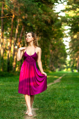 Obraz na płótnie Canvas Beautiful curly hair young woman wearing elegant purple dress standing on a path in the middle of forest with rays of sunlight during sunset