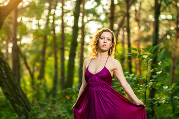 Beautiful curly hair young woman wearing elegant purple dress standing in the middle of forest with rays of sunlight during sunset