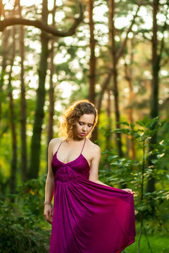 Beautiful curly hair young woman wearing elegant purple dress standing in the middle of forest with rays of sunlight during sunset