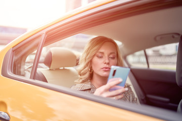 Photo of blonde with phone in her hand sitting in back seat in yellow taxi in afternoon