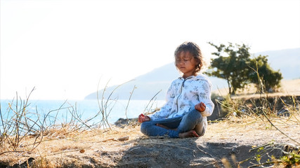 Cute little child gurl meditating alone in lotus pose at lake shore in windy weather