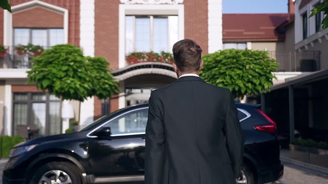 Slow motion of slender handsome businessman walking and sitting to the black car standing near beautiful house