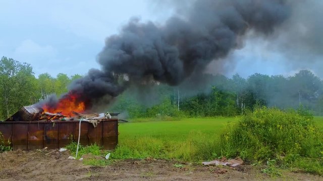 Thick dark smoke billowing from a burning dumpster full of garbage on rural farmland.
