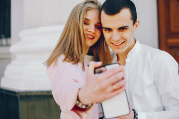 beautiful and stylish blonde along with her handsome guy standing near building in the summer city and use the phone in white case