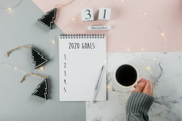 Happy New Year 2020. Woman's hand holding cup of coffee and writing 2020 Goals in notebook...