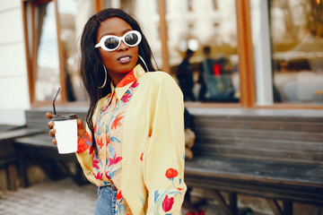 Fototapeta premium A beautiful and stylish black girl with long dark hair dressed in a yellow shirt and with sunglasses standing a summer city with coffee