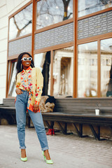 A beautiful and stylish black girl with long dark hair dressed in a yellow shirt and with sunglasses standing in a summer city