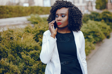 elegant and stylish dark-skinned girl with curly hair and in a white jacket walking around the summer city and using the phone