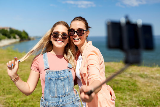 leisure and friendship concept - happy smiling teenage girls or friends in sunglasses hugging and taking picture by smartphone on selfie stick at seaside in summer