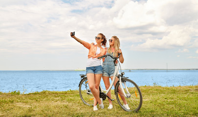 leisure, technology and friendship concept - happy smiling teenage girls or friends with bicycle taking selfie by smartphone at seaside in summer