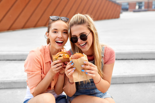 leisure, fast food and friendship concept - happy smiling teenage girls or best friends in sunglasses eating burgers on city street in summer