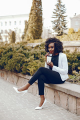 elegant and stylish dark-skinned girl with curly hair and in a white jacket sitting in the summer city near green brush and drinking a coffee