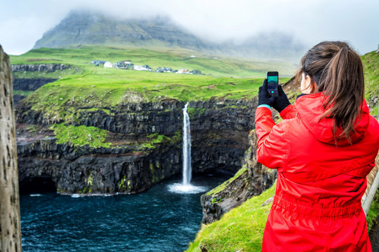 Young female travelers is taking photo of Mulafossur Waterfall with the village Gasadalur in background, Faroe Islands.
