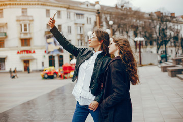 Obraz premium a beautiful stylish young girl with long curly hair and a long coat walking in the autumn city with her girlfriend in a black leather jacket and glasses and they uses the phone