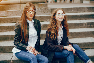 a beautiful stylish young girl with long curly hair and a long coat sitting on a stairs in the autumn city with her girlfriend in a black leather jacket and glasses