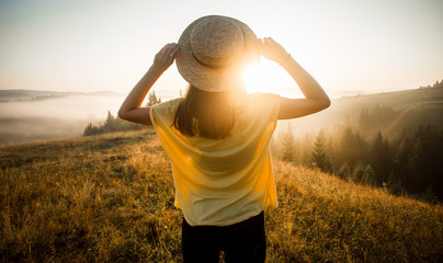 Back view of woman in straw hat stay outdoor under sunlight of sunrise with beautiful view of forest mountains - obrazy, fototapety, plakaty
