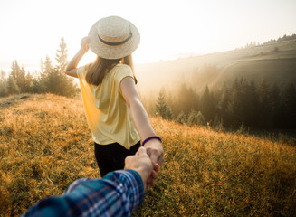 Follow me concept. Couple in love holding hands. Woman in yellow shirt and straw hat holding man by hand going to autumn forest with mountains and cloudy sky in morning. Woman leads man.