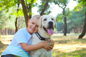 Portrait of cute blond boy hanging out with his pet friend labrador retriever out in the woods. Little guy hugging six months old doggy, walk in a park. Background, copy space, close up.