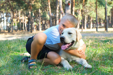 Portrait of cute blond boy hanging out with his pet friend labrador retriever out in the woods. Little guy hugging six months old doggy, walk in a park. Background, copy space, close up.