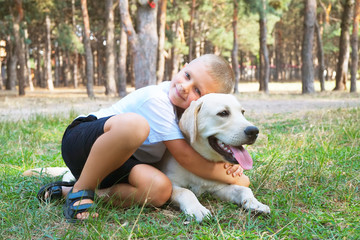 Portrait of cute blond boy hanging out with his pet friend labrador retriever out in the woods. Little guy hugging six months old doggy, walk in a park. Background, copy space, close up.