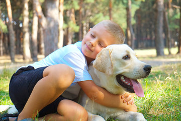 Portrait of cute blond boy hanging out with his pet friend labrador retriever out in the woods. Little guy hugging six months old doggy, walk in a park. Background, copy space, close up.