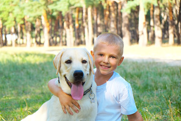 Portrait of cute blond boy hanging out with his pet friend labrador retriever out in the woods. Little guy hugging six months old doggy, walk in a park. Background, copy space, close up.