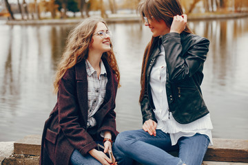 a beautiful stylish young girl with long curly hair and a long coat sitting in the autumn park near river with her girlfriend in a black leather jacket and glasses and they use the phone