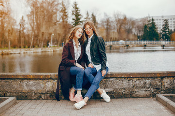 a beautiful stylish young girl with long curly hair and a long coat sitting in the autumn park near river with her girlfriend in a black leather jacket and glasses