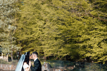 Elegant bride and groom posing together outdoors on a wedding day. Newlywed couple with roses bouquet posing and kissing. Happy newlyweds posing in the park on their wedding day. 