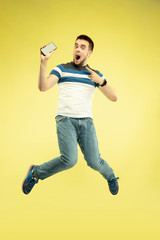 Fototapeta na wymiar Communication. Full length portrait of happy jumping man with gadgets on yellow background. Modern tech, freedom of choices concept, emotions concept. Using smartphone for selfie or videocall in
