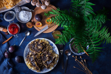 Obraz na płótnie Canvas Sweet pasta dessert, noodles with poppy seeds, plum compote from fresh plums, dark background, blue table decorated with fresh flowers. Dry whole poppy plant in background.