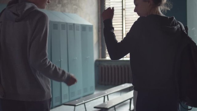 Back view of two female friends coming to lock room, greeting each other by bumping their fists then putting their backpacks on bench