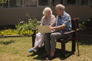 Front view of senior couple sitting on bench and using laptop in garden