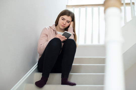 Unhappy Teenage Girl Being Bullied By Text Message Sitting On Stairs At Home