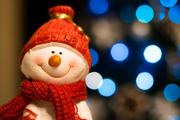 Happy smiling snowman wearing red scarf and hat with Christmas tree and Christmas lights in the background.