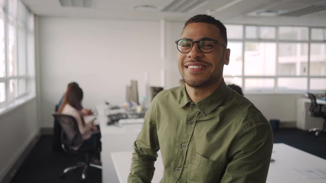 Portrait of successful professional young african american businessman smiling and looking at camera sitting in front of colleague working at background