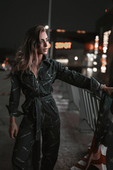 Street portrait of stylish fashion girl in denim overalls on parking at night. Young woman walking in evening in neon light near shops with beautiful hair style