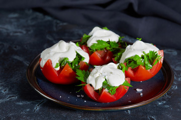 Close - up of tomato halves with finely chopped parsley in a sauce of sour cream and salt lie on a plate on a dark background. Very tasty vegetarian salad.