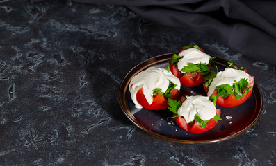 Close - up of tomato halves with finely chopped parsley in a sauce of sour cream and salt lie on a plate on a dark background. Horizontal image. Copy space.