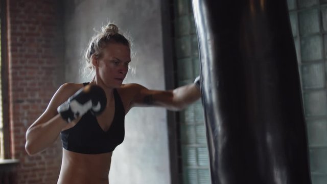 Medium shot of female Caucasian boxer with blonde hair wearing gloves hitting big black punching bag