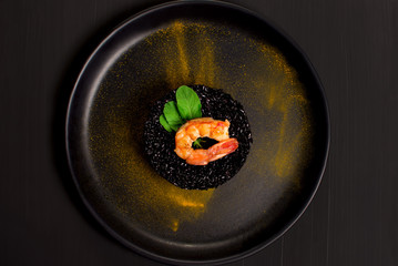 Black rice risotto with shrimp and safron in a black plate seen from above in dark background