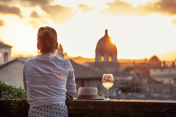 Young woman tourist fashion white dress with glass of white wine in front of panoramic view of Rome cityscape from campidoglio terrace at sunset. Landmarks and domes. - obrazy, fototapety, plakaty