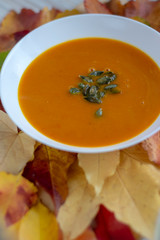 Tasty pumpkin bright orange soup with green seeds served on white plate and autumnal colorful leaves