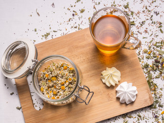 Composition with dried chamomile, glass mug with hot tea, meringues, bamboo board and dried herbal leaves and petals