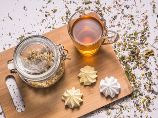Composition with dried chamomile, glass mug with hot tea, meringues, bamboo board and dried herbal leaves and petals