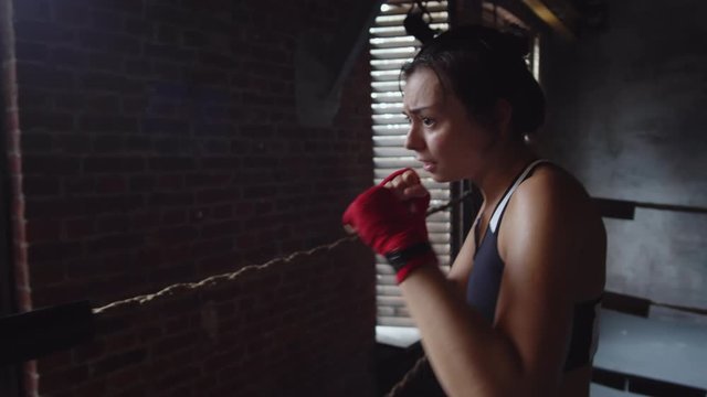 Medium shot of young female athlete wearing racerback tank top, shorts and red bandages on hands doing multiple punches inside boxing ring then looking at camera