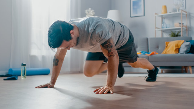 Smiling Strong Athletic Fit Man in T-shirt and Shorts is Doing Mountain Climber Exercises at Home in His Spacious and Bright Living Room with Minimalistic Interior.