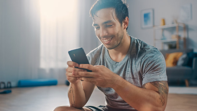 Happy Strong Athletic Fit Man in T-shirt and Shorts is Using a Mobile Phone After Morning Exercises at Home in His Spacious and Bright Living Room with Minimalistic Interior.